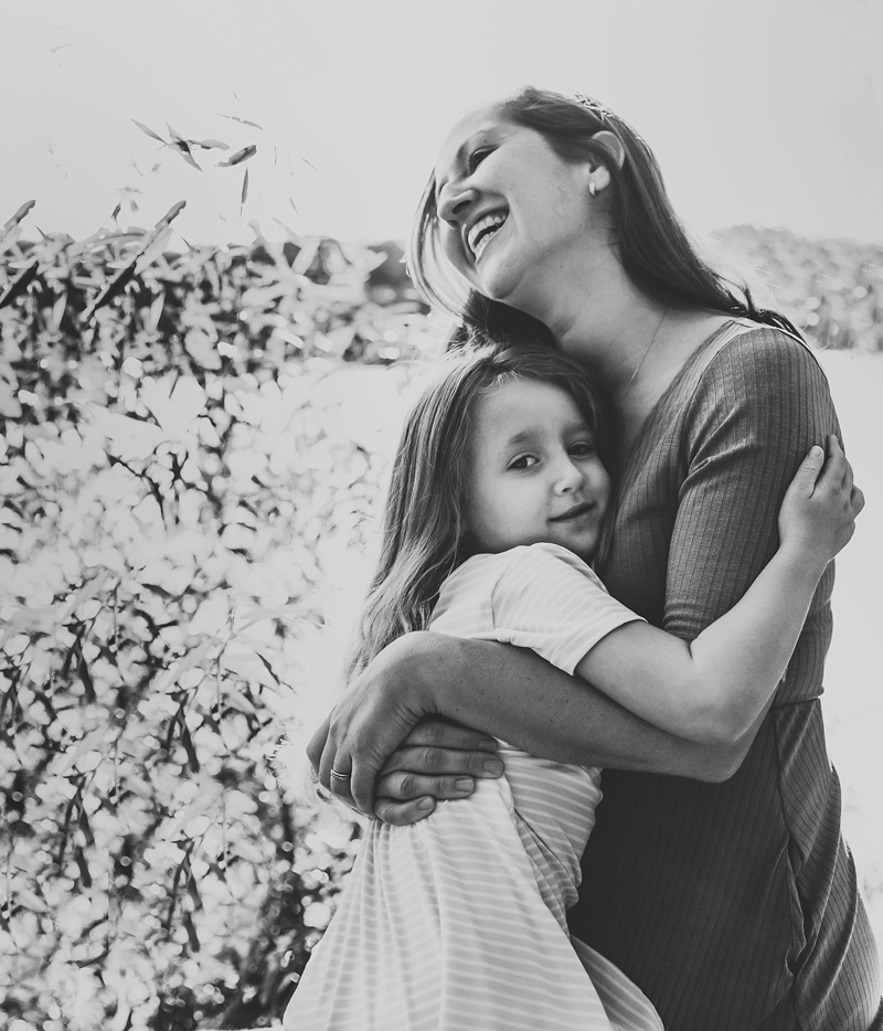 family photo, mother laughs as she squeezes happy daughter outside in a field