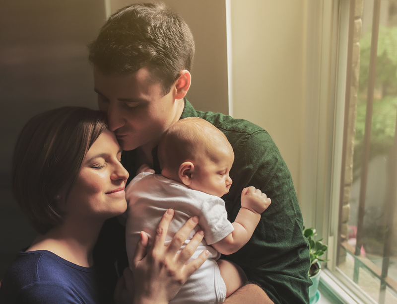 Family photography, husband embraces wife while holding newborn baby near window