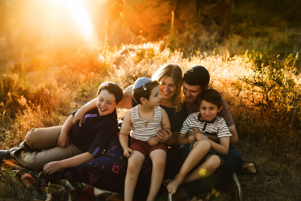 Family Photographer, Chana Grossman, sits holding her husband and three boys outside at golden hour